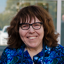 Nancy Ward smiles, wearing a blue and green floral shirt. She has brown hair and black glasses.